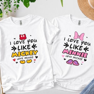 Pode incluir: Duas camisetas brancas com gr&aacute;ficos correspondentes. Uma camisa diz "I love you like Mickey" com um la&ccedil;o vermelho e detalhes do Mickey Mouse. A outra diz "I love you like Minnie" com um la&ccedil;o rosa e detalhes da Minnie Mouse.