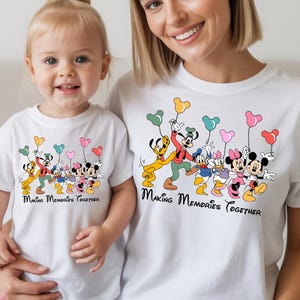 May include: White t-shirts featuring a colorful graphic of Disney characters holding balloon bouquets. The text "Making Memories Together" is printed below the characters. The shirts are worn by a mother and child.