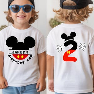 May include: White t-shirt featuring a black Mickey Mouse head design with the name "Jakson" and the words "Birthday Boy" on the front. The back of the shirt displays a large number "2" in black and red, with Mickey Mouse hands on either side.