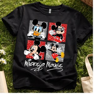 Op de afbeelding: Zwart T-shirt met vier panelen met Mickey Mouse in verschillende poses. De panelen hebben rode en zilveren glitterachtergronden. De woorden "Mickey Mouse" staan eronder in een cursief lettertype.