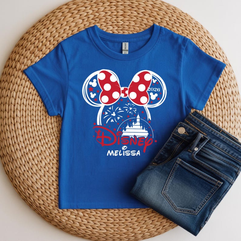 May include: A royal blue t-shirt with a white and red graphic of a mouse head with a polka dot bow, fireworks, and a castle. The word "Disney" is in red, with the name "Melissa" below. A pair of blue jeans is next to the shirt.