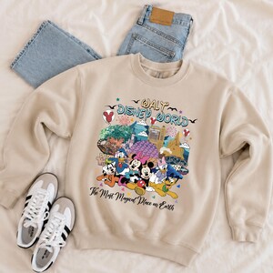 K&ouml;nnte beinhalten: Beiges Sweatshirt mit Rundhalsausschnitt und farbenfroher Grafik mit Disney-Figuren und Wahrzeichen. Der Text "Walt Disney World" und "The Most Magical Place on Earth" ist auf der Vorderseite aufgedruckt. Enth&auml;lt ein Paar wei&szlig;e Turnschuhe und blaue Jeans.