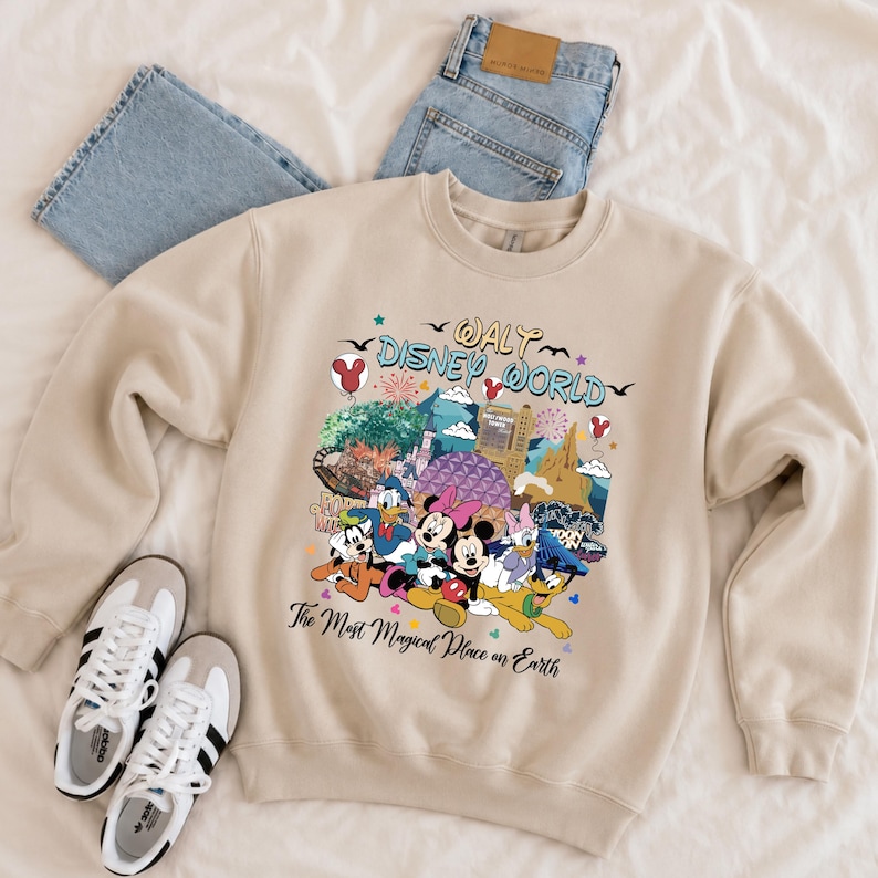 K&ouml;nnte beinhalten: Beiges Sweatshirt mit Rundhalsausschnitt und farbenfroher Grafik mit Disney-Figuren und Wahrzeichen. Der Text "Walt Disney World" und "The Most Magical Place on Earth" ist auf der Vorderseite aufgedruckt. Enth&auml;lt ein Paar wei&szlig;e Turnschuhe und blaue Jeans.