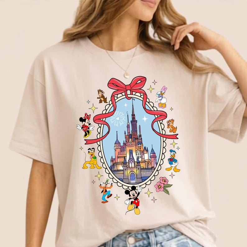May include: Beige t-shirt featuring a colorful Disney castle design within an oval frame, surrounded by various Disney characters. The design includes Mickey Mouse, Minnie Mouse, Donald Duck, and others, with a red bow at the top.
