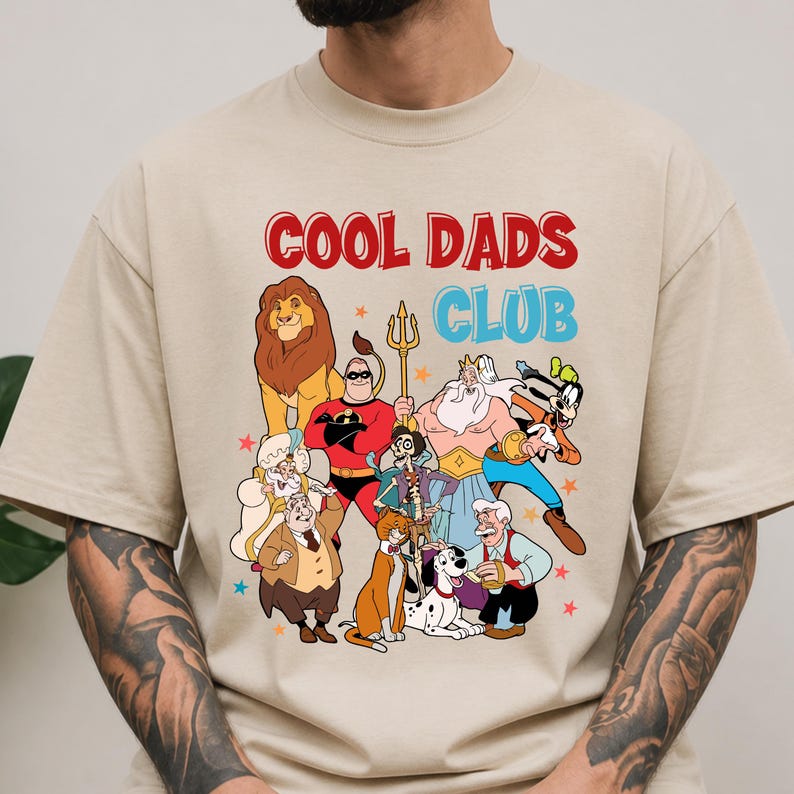 Pode incluir: Camiseta bege com o texto "COOL DADS CLUB" em vermelho e azul. A camiseta apresenta personagens de desenhos animados, incluindo um le&atilde;o, um super-her&oacute;i e outras figuras animadas. O design est&aacute; centrado na frente da camiseta.