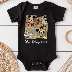 Puede incluir: Body de beb&eacute; negro con cuatro paneles cuadrados con personajes de Disney con trajes de safari. Se representan Mickey Mouse, Minnie Mouse, Goofy y Donald Duck. Las palabras "Walt Disney World" est&aacute;n impresas en blanco.