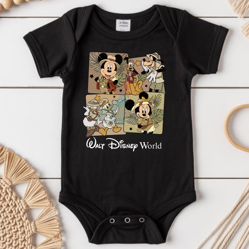 Puede incluir: Body de beb&eacute; negro con cuatro paneles cuadrados con personajes de Disney con trajes de safari. Se representan Mickey Mouse, Minnie Mouse, Goofy y Donald Duck. Las palabras "Walt Disney World" est&aacute;n impresas en blanco.
