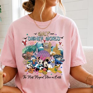 K&ouml;nnte beinhalten: Hellrosa T-Shirt mit einem farbenfrohen Aufdruck von Disney-Figuren und Wahrzeichen, darunter Micky und Minnie Maus, Donald Duck und das Cinderella-Schloss. Der Text "Walt Disney World" und "The Most Magical Place on Earth" sind ebenfalls abgebildet.