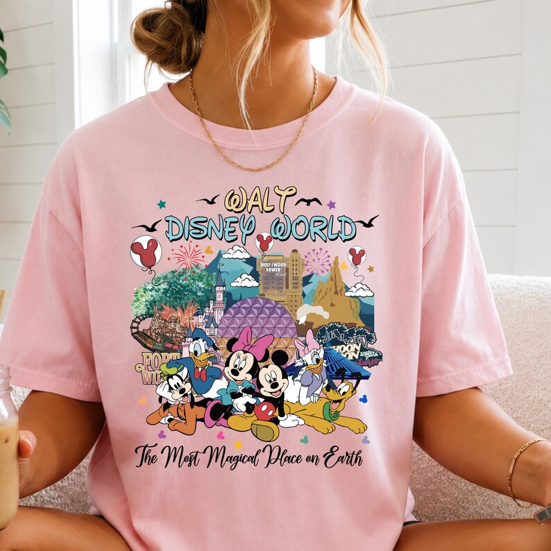 K&ouml;nnte beinhalten: Hellrosa T-Shirt mit einem farbenfrohen Aufdruck von Disney-Figuren und Wahrzeichen, darunter Micky und Minnie Maus, Donald Duck und das Cinderella-Schloss. Der Text "Walt Disney World" und "The Most Magical Place on Earth" sind ebenfalls abgebildet.