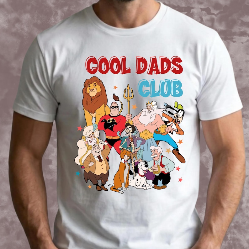 Pode incluir: Camiseta branca com o texto "COOL DADS CLUB" em vermelho e azul. A camisa apresenta personagens de desenhos animados, incluindo um le&atilde;o, um super-her&oacute;i e outros. Estrelas coloridas est&atilde;o espalhadas ao redor dos personagens.