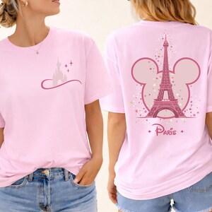 May include: Light pink t-shirt featuring a castle graphic on the front and the Eiffel Tower with a Mickey Mouse ear outline on the back. The word "Paris" is printed below the tower. The shirt is a soft, casual style.