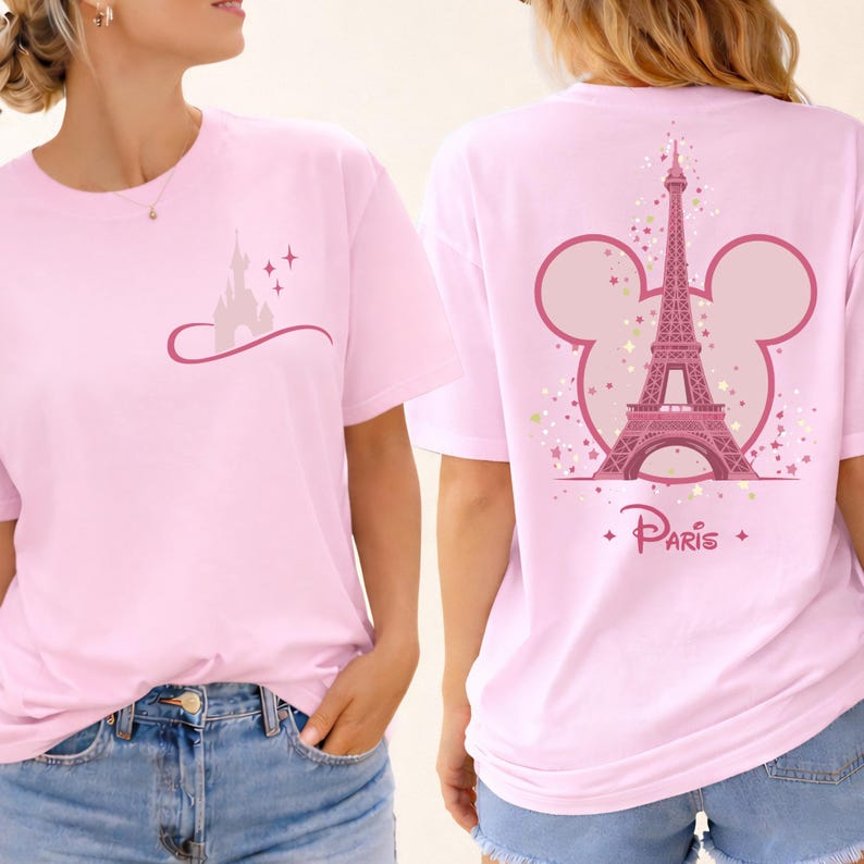 May include: Light pink t-shirt featuring a castle graphic on the front and the Eiffel Tower with a Mickey Mouse ear outline on the back. The word "Paris" is printed below the tower. The shirt is a soft, casual style.