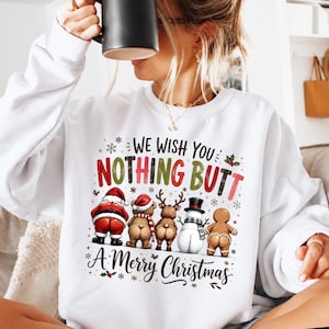 May include: A white long-sleeve sweatshirt featuring a graphic print with the text "WE WISH YOU NOTHING BUTT A MERRY CHRISTMAS" and illustrations of Santa, a bear, a reindeer, a snowman, and a gingerbread person all facing away.