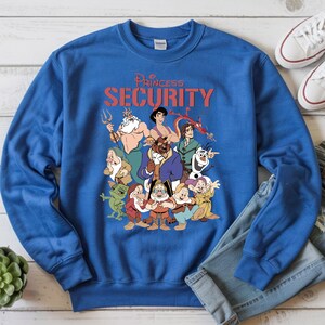 May include: Royal blue sweatshirt with a graphic of Disney characters and the text "Princess Security" in red. The design includes characters such as King Triton, Aladdin, Beast, and the Seven Dwarfs. White trainers and blue jeans are also visible.