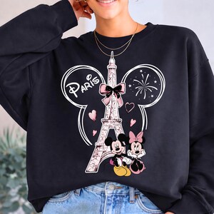K&ouml;nnte beinhalten: Schwarzes Sweatshirt mit einem Aufdruck des Eiffelturms mit rosa Schleife, dem Wort "Paris" und Mickey und Minnie Maus. Das Design umfasst rosa Herzen und Feuerwerk, wodurch ein verspieltes Thema entsteht.