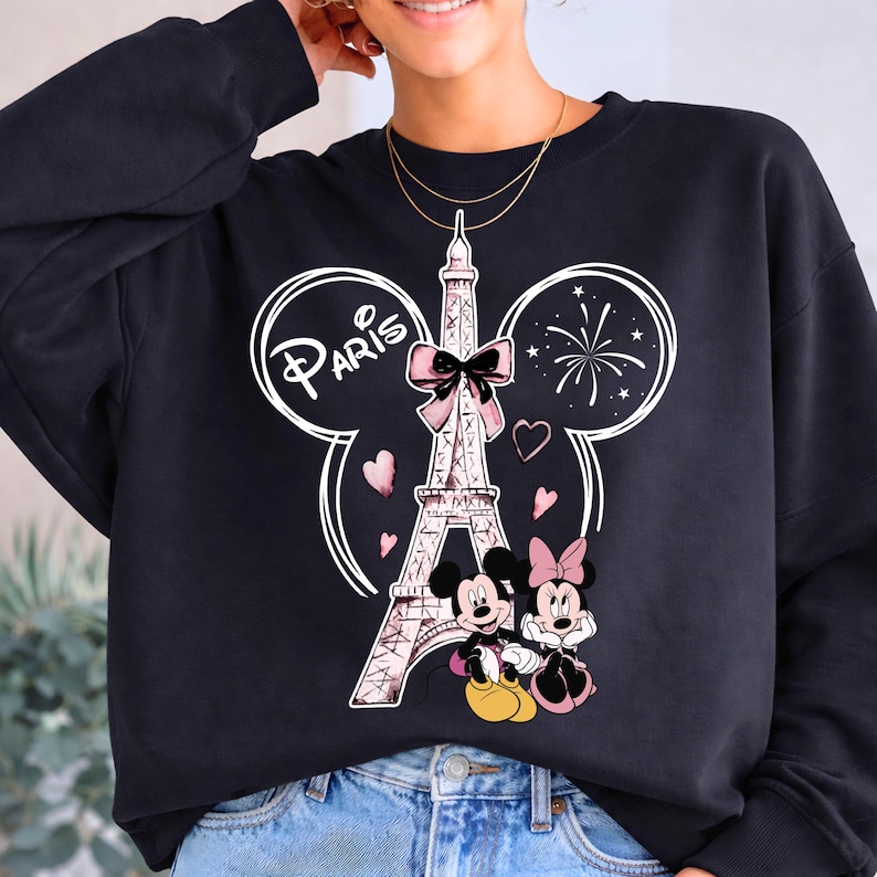 K&ouml;nnte beinhalten: Schwarzes Sweatshirt mit einem Aufdruck des Eiffelturms mit rosa Schleife, dem Wort "Paris" und Mickey und Minnie Maus. Das Design umfasst rosa Herzen und Feuerwerk, wodurch ein verspieltes Thema entsteht.