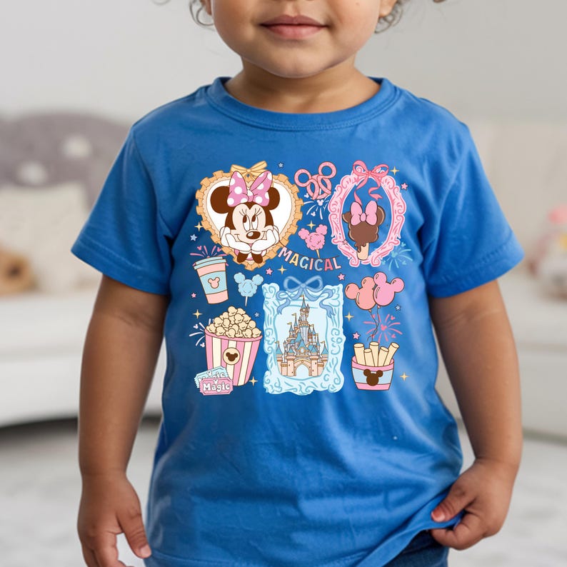 Puede incluir: Camiseta azul con un gr&aacute;fico colorido que presenta a Minnie Mouse, palomitas de ma&iacute;z, un castillo y la palabra "MAGICAL". El dise&ntilde;o incluye detalles en rosa y dorado, creando una est&eacute;tica l&uacute;dica y caprichosa. La camiseta es de material suave.