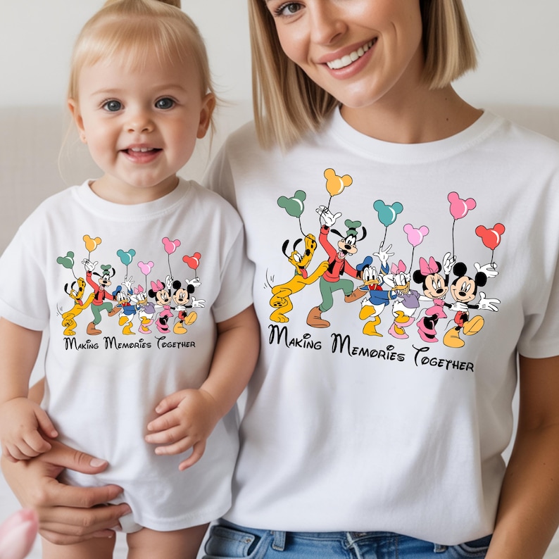 May include: White t-shirts featuring a colorful graphic of Disney characters holding balloon bouquets. The text "Making Memories Together" is printed below the characters. The shirts are worn by a mother and child.