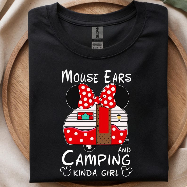 Puede incluir: Camiseta negra con un gr&aacute;fico de una caravana dise&ntilde;ada como orejas y lazo de Minnie Mouse. El texto dice "Mouse Ears and Camping Kinda Girl". La caravana tiene lunares rojos y una puerta roja.