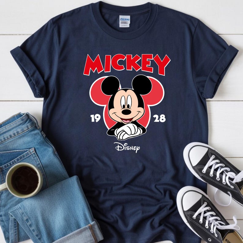 May include: Navy blue t-shirt featuring a Mickey Mouse graphic. The design includes the word "MICKEY" in red, the numbers "19" and "28", and the Disney logo. The shirt is paired with blue jeans and black sneakers.