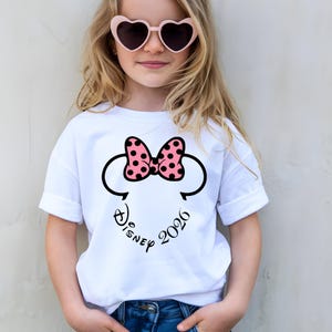 May include: White t-shirt featuring a Minnie Mouse design with a pink polka-dot bow and black outlines. The text "Disney 2026" is printed below the bow. The shirt is worn by a child wearing heart-shaped sunglasses.