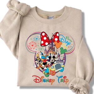 Puede incluir: Sudadera beige con un gr&aacute;fico colorido de personajes de Disney en un coche, con un castillo, fuegos artificiales y un lazo de Minnie Mouse. Tambi&eacute;n se muestran las palabras "Best Day" y "Disney Trip".