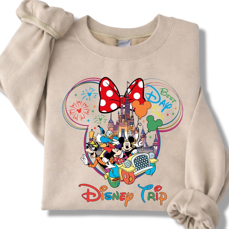 Puede incluir: Sudadera beige con un gr&aacute;fico colorido de personajes de Disney en un coche, con un castillo, fuegos artificiales y un lazo de Minnie Mouse. Tambi&eacute;n se muestran las palabras "Best Day" y "Disney Trip".