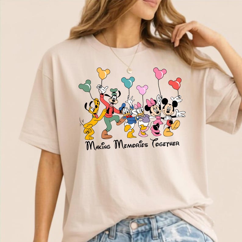 May include: Beige t-shirt featuring a colorful graphic of Disney characters, including Mickey and Minnie Mouse, Goofy, Donald and Daisy Duck, and Pluto, holding balloons. The text "Making Memories Together" is printed below the characters.