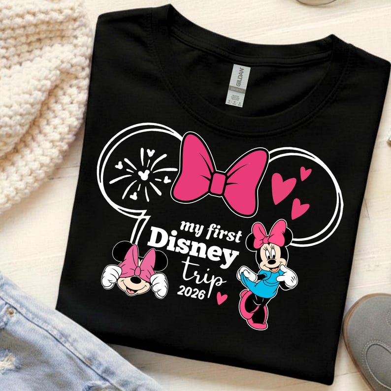 May include: Black t-shirt featuring a Minnie Mouse design with a pink bow, hearts, and the text "my first Disney trip 2026". The design includes Minnie Mouse illustrations and fireworks.