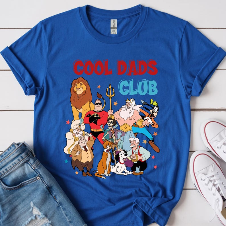 Pode incluir: Uma camiseta azul royal com as palavras "COOL DADS CLUB" em vermelho e azul, com personagens de desenhos animados. A camiseta &eacute; combinada com jeans azuis e t&ecirc;nis brancos.