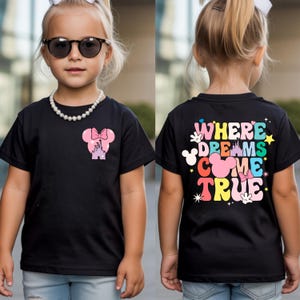 May include: Black t-shirt with a Minnie Mouse design on the front and the text "WHERE DREAMS COME TRUE" on the back. The design includes a pink bow, castle, and colourful text. The shirt is worn by a child.