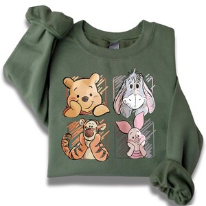 May include: Olive green sweatshirt featuring four cartoon character portraits: Winnie the Pooh, Eeyore, Tigger, and Piglet. Each character is in a separate square, with a sketched style. The sweatshirt has long sleeves and a crew neck.