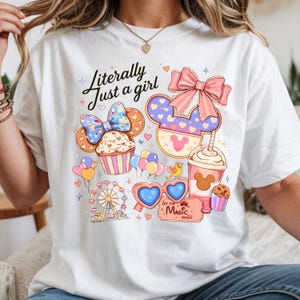Pode incluir: Camiseta branca com o texto "Literally Just a girl" e um gr&aacute;fico colorido com um cupcake, orelhas do Mickey, uma roda gigante, &oacute;culos de sol em forma de cora&ccedil;&atilde;o e uma x&iacute;cara de caf&eacute;. O gr&aacute;fico inclui detalhes em rosa, azul e amarelo.