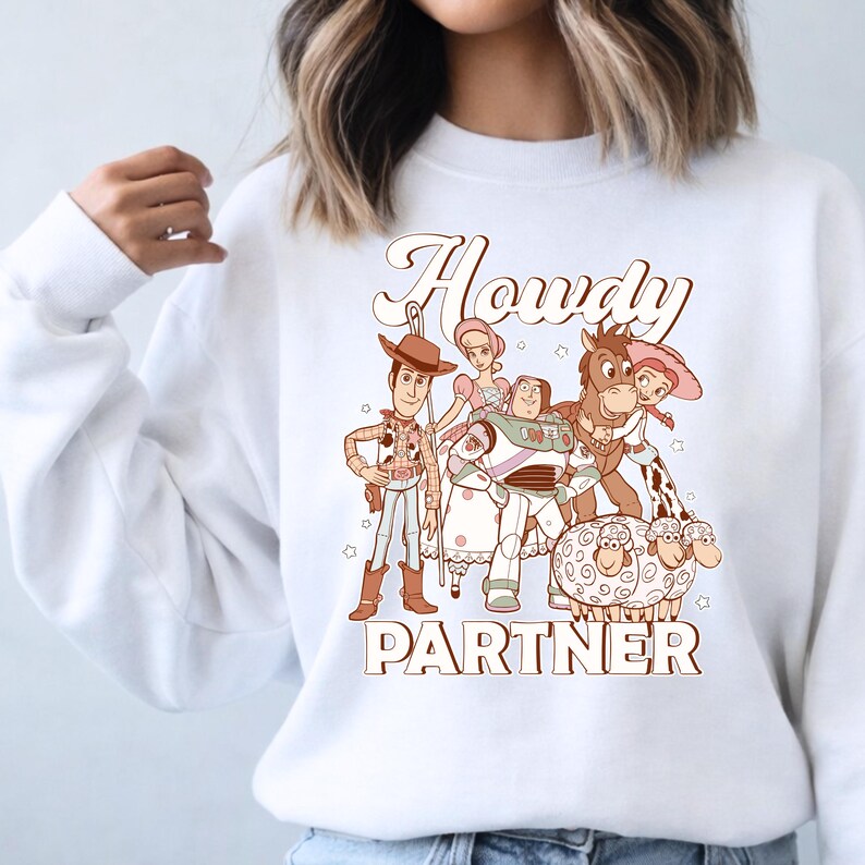 May include: White sweatshirt featuring a cartoon graphic of characters from the movie Toy Story. The graphic includes Woody, Buzz Lightyear, Jessie, and other characters, with the words "Howdy Partner" in a brown font.