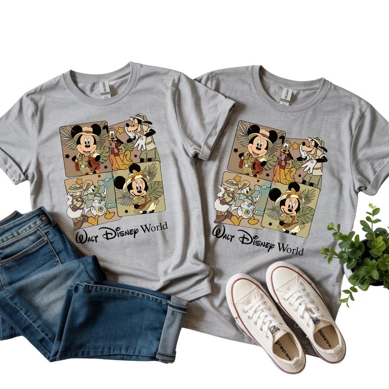 Puede incluir: Dos camisetas grises con un gr&aacute;fico colorido con Mickey Mouse, el Pato Donald, Goofy y otros personajes de Disney con trajes de safari. El texto "Walt Disney World" est&aacute; impreso debajo. Tambi&eacute;n hay unos vaqueros azules y zapatillas blancas.