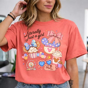 Pode incluir: Camiseta coral com um gr&aacute;fico l&uacute;dico. Apresenta um cupcake, orelhas do Mickey Mouse, uma roda gigante, &oacute;culos de sol em forma de cora&ccedil;&atilde;o e o texto "Literally Just a girl". O design inclui la&ccedil;os, bal&otilde;es e uma x&iacute;cara de caf&eacute;.