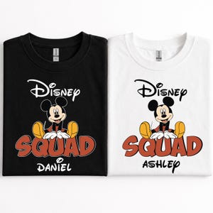 Op de afbeelding: Twee T-shirts, &eacute;&eacute;n zwart en &eacute;&eacute;n wit, met een Mickey Mouse-afbeelding. De shirts tonen het woord "Disney" boven het personage en "SQUAD" in rode letters. De naam "DANIEL" staat op het zwarte shirt en "ASHLEY" op het witte.