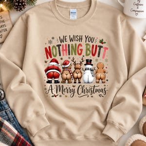 May include: A beige crewneck sweatshirt featuring a graphic of Santa, a bear, a reindeer, a snowman, and a gingerbread person facing away. The text reads "WE WISH YOU NOTHING BUTT A MERRY CHRISTMAS" in red, green, and black lettering with snowflakes.