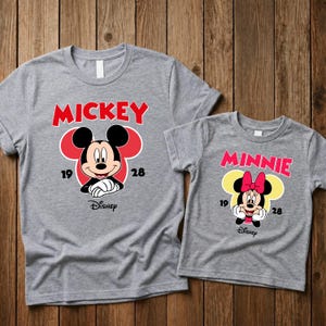 May include: Two gray t-shirts with Disney character designs. One shirt features "Mickey" in red text above a Mickey Mouse graphic, the other features "Minnie" above a Minnie Mouse graphic. Both shirts have the Disney logo.