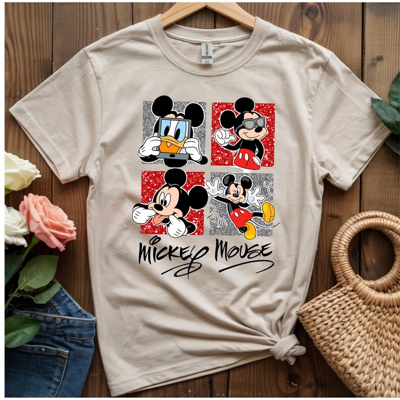 Op de afbeelding: Een beige t-shirt met een afbeelding van Mickey Mouse in vier verschillende poses. Elke pose staat in een vierkant, met de woorden "Mickey Mouse" in een cursief lettertype eronder. Het shirt hangt aan een houten hanger.