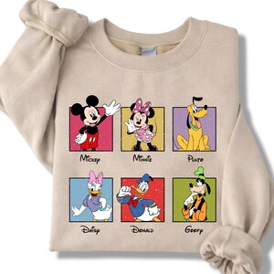 May include: Beige sweatshirt featuring six Disney characters: Mickey, Minnie, Pluto, Daisy, Donald, and Goofy. Each character is in a colorful square with their name below. The sweatshirt has long sleeves.