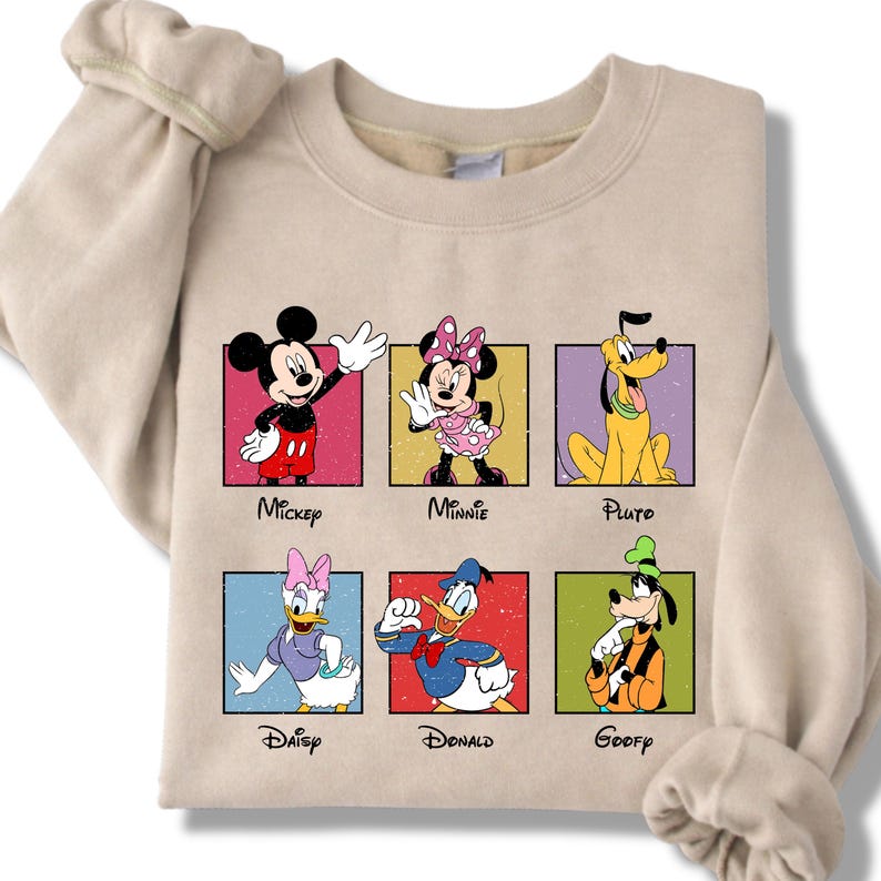 May include: Beige sweatshirt featuring six Disney characters: Mickey, Minnie, Pluto, Daisy, Donald, and Goofy. Each character is in a colorful square with their name below. The sweatshirt has long sleeves.