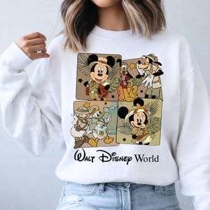 Puede incluir: Sudadera blanca con cuatro paneles cuadrados con personajes de Disney con trajes de safari. Se representan Mickey Mouse, Pluto, Goofy, Donald y Minnie Mouse. Las palabras "Walt Disney World" est&aacute;n impresas en negro debajo.