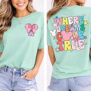 May include: A pale green t-shirt with a vibrant graphic on the front and back. The front displays a pink Mickey Mouse head with a bow and a castle. The back says "Where Dreams Come True" in colourful letters.