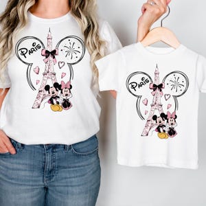 K&ouml;nnte beinhalten: Wei&szlig;e T-Shirts mit einem Design mit dem Eiffelturm, Mickey und Minnie Mouse und dem Wort "Paris". Das Design umfasst rosa Schleifen, Herzen und Feuerwerk. Ein T-Shirt h&auml;ngt an einem Kleiderb&uuml;gel, das andere wird getragen.