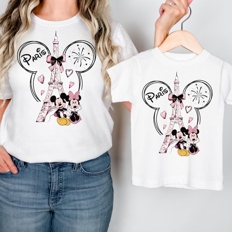 K&ouml;nnte beinhalten: Wei&szlig;e T-Shirts mit einem Design mit dem Eiffelturm, Mickey und Minnie Mouse und dem Wort "Paris". Das Design umfasst rosa Schleifen, Herzen und Feuerwerk. Ein T-Shirt h&auml;ngt an einem Kleiderb&uuml;gel, das andere wird getragen.