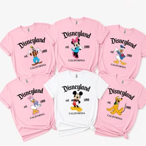 Op de afbeelding: Een verzameling roze en witte t-shirts met Disney-personages. Elk shirt toont het Disneyland-logo, het jaar 1955 en CALIFORNIA. Personages zijn onder meer Mickey, Minnie, Donald, Daisy, Goofy en Pluto.