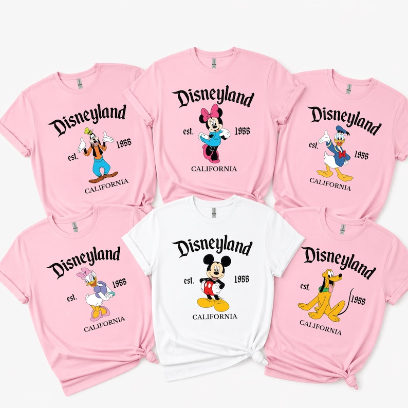 Op de afbeelding: Een verzameling roze en witte t-shirts met Disney-personages. Elk shirt toont het Disneyland-logo, het jaar 1955 en CALIFORNIA. Personages zijn onder meer Mickey, Minnie, Donald, Daisy, Goofy en Pluto.