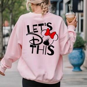 K&ouml;nnte beinhalten: Hellrosa Sweatshirt mit dem schwarzen Text "LET'S DO THIS" und einem Minnie Mouse-Motiv mit roter Schleife und Schloss-Silhouette. Das Sweatshirt hat lange &Auml;rmel.