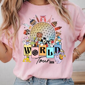 May include: Pale pink sweatshirt with a vibrant graphic of Disney characters and global landmarks. The design includes Mickey Mouse, Donald Duck, and the text "WORLD Tour 2026". The sweatshirt has long sleeves.